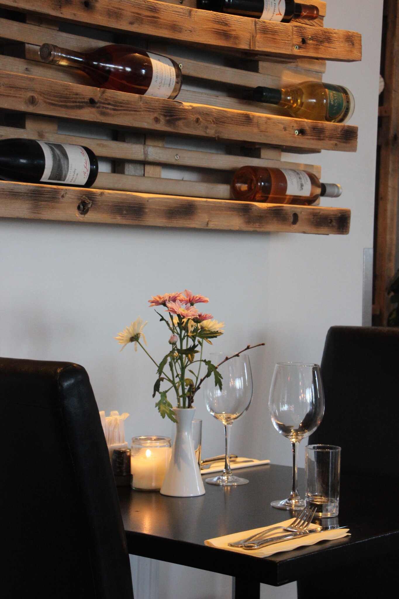 Et hyggeligt restaurantmiljø med et lille bord til to, med vinglas, en hvid vase med blomster, bestik og et stearinlys. Træstativer med vinflasker er monteret på væggen, hvilket forstærker den intime atmosfære.