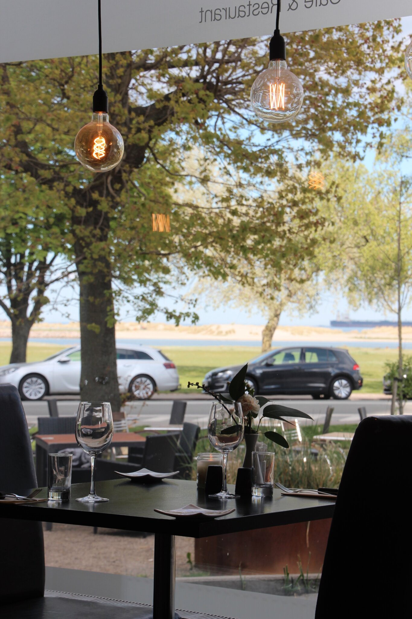Et hyggeligt restaurantinteriør med et bord til to, med vinglas og en lille blomstervase. Store vinduer afslører en udsigt over parkerede biler, træer og en fjern vandmasse under en klar, solrig himmel. Hængende pærer tilføjer en varm atmosfære.