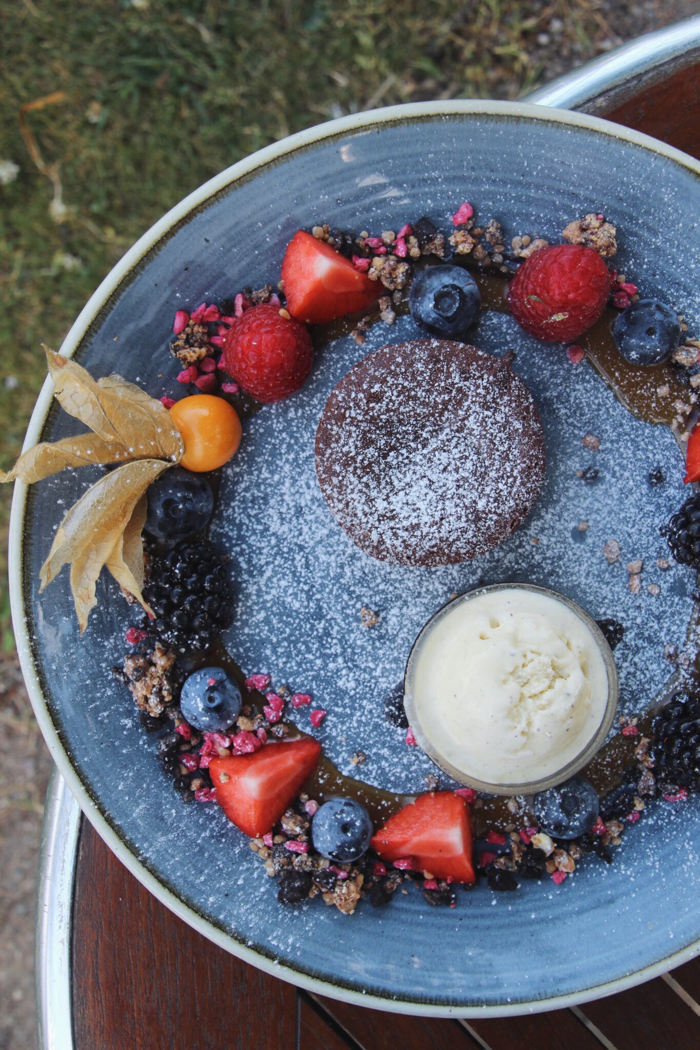 En desserttallerken har en smeltet chokoladekage, der er pudret med pulveriseret sukker, omgivet af friske jordbær, hindbær, blåbær og physalis. En kugle vaniljeis er også på tallerkenen, pyntet med granolastumper og granatæblekerner.