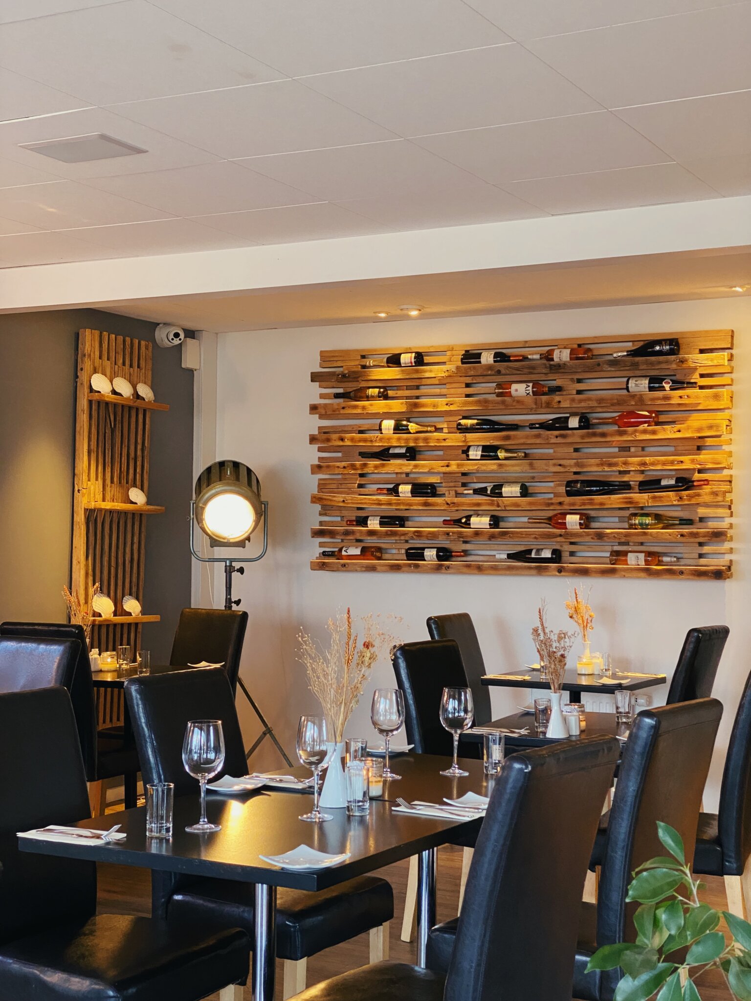 Et hyggeligt restaurantinteriør med sorte læderstole og mørke træborde. Vinflasker er udstillet på en vægreol i træ. Bordene er dækket med tomme vinglas og minimalistiske tørrede blomsterarrangementer. En vintage gulvlampe tilføjer omgivende lys.