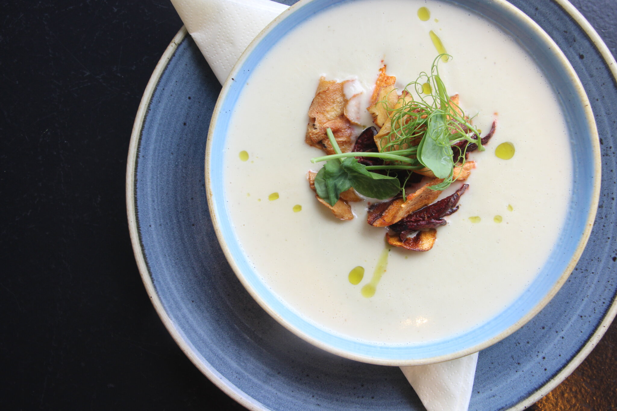 En skål cremet suppe pyntet med krydderurter, små kødstykker og et skvæt olie står på en blå tallerken med en hvid serviet. Indstillingen er på en mørk overflade, hvilket skaber en kontrast til den lyse suppe.