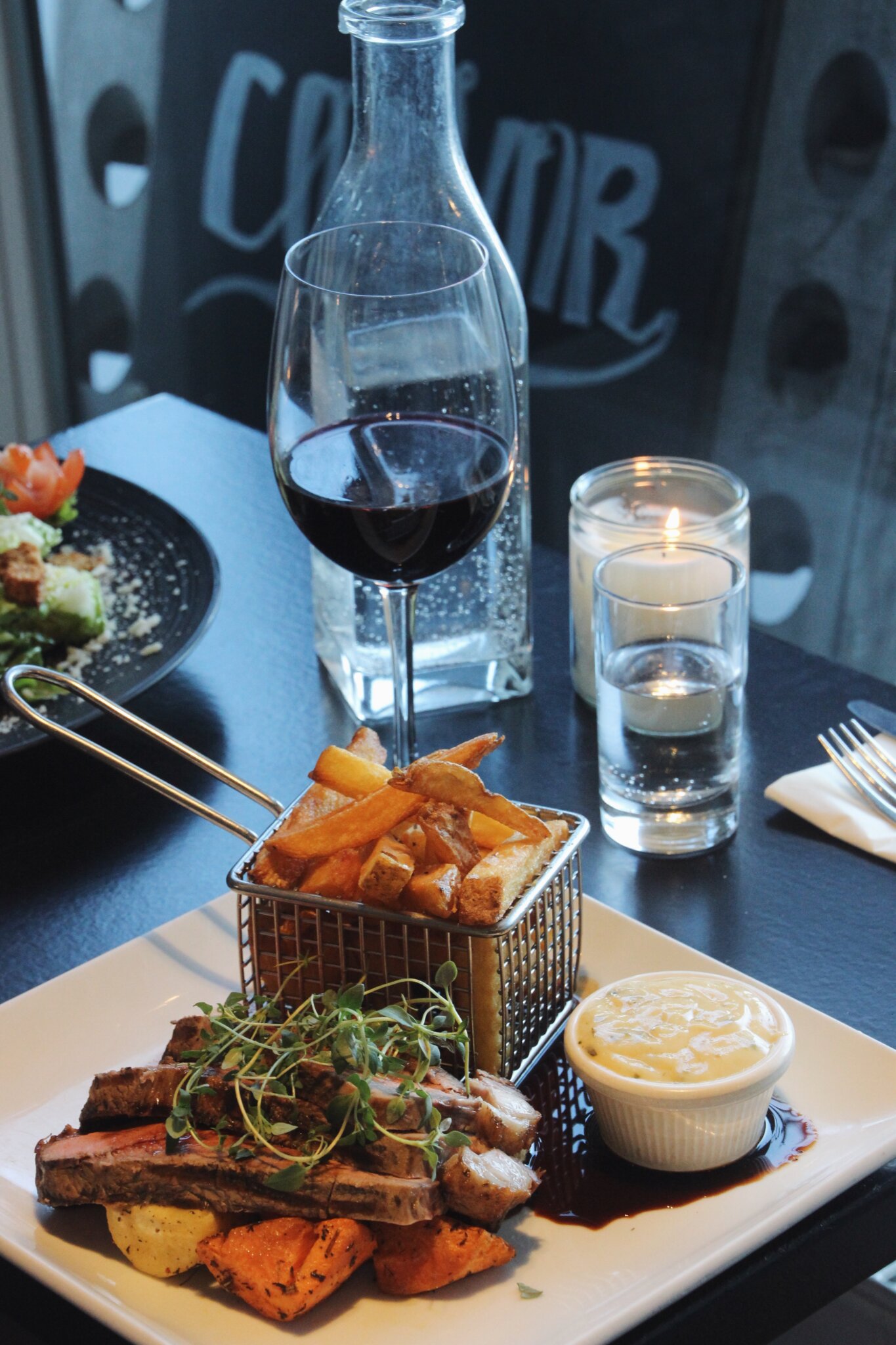 Et spisebord med et glas rødvin, en tallerken bøf toppet med grønt, søde kartoffeltern og et lille fad sauce. En kurv med fritter ledsager måltidet. Et stearinlys, et glas vand og en serviet med bestik står også på bordet.