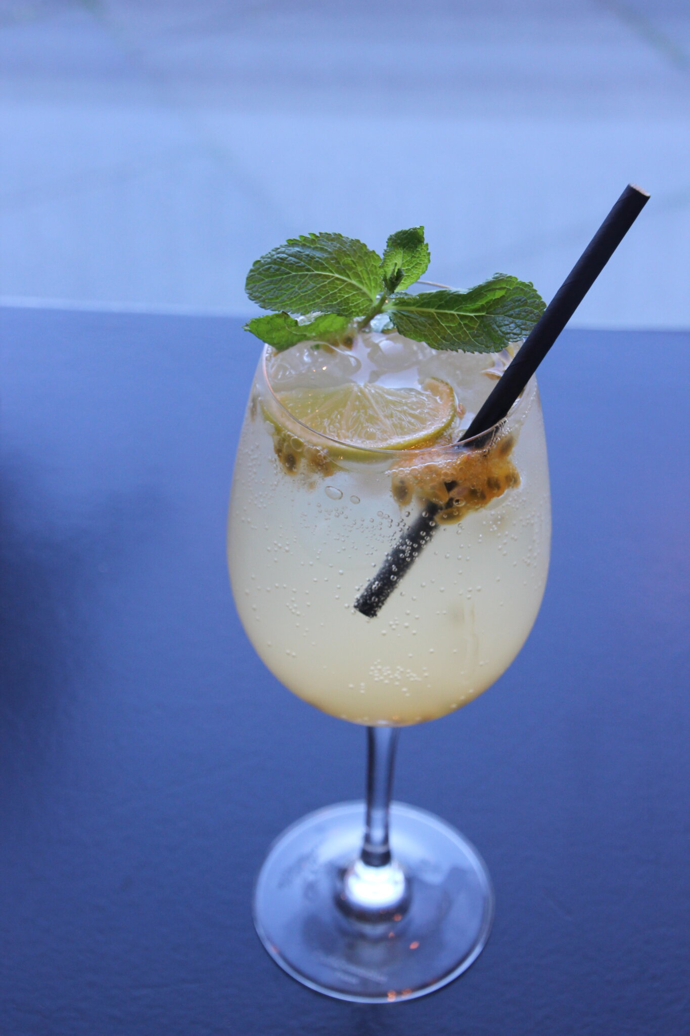En forfriskende cocktail i et vinglas med et sort sugerør, garneret med limeskiver, mynteblade og passionsfrugtfrø, sat mod en sløret blå baggrund.