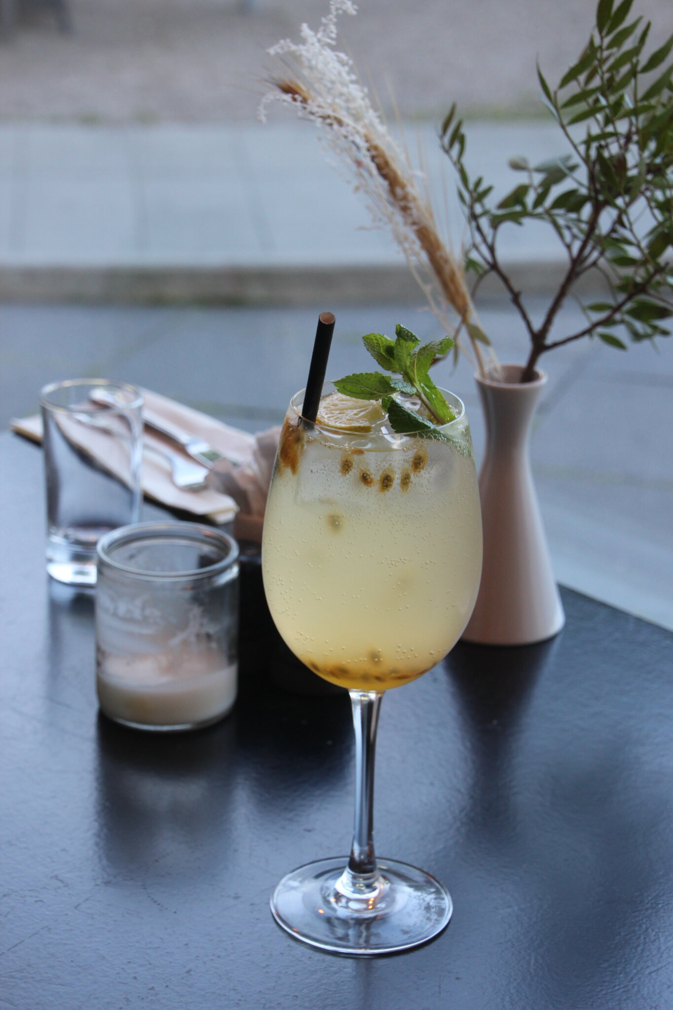 En forfriskende cocktail i et vinglas garneret med mynte og skiver af lime står på et mørkt bord. I nærheden er et lille lys, to tomme glas og en vase med tørt og grønt løv, med sløret udendørs landskab i baggrunden.