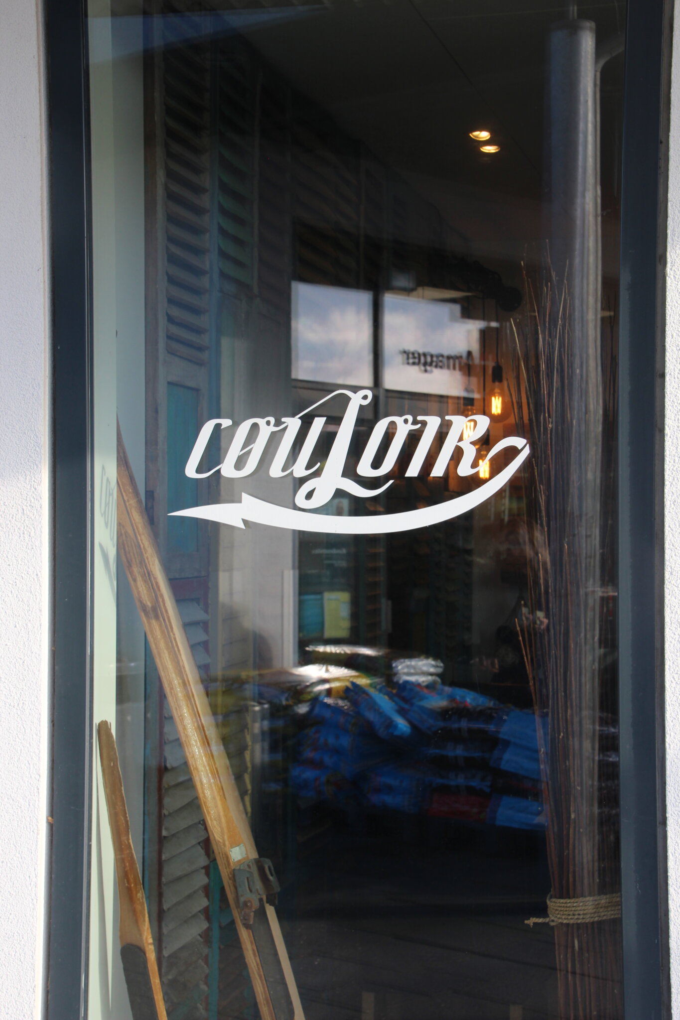 En glaslåge med ordet COULOIR i stiliserede hvide bogstaver. Refleksion af indvendige lys og en stak genstande er synlig. Ski og høje dekorative pinde er placeret nær døren.